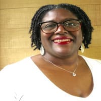 Smiling Black woman wearing glasses and a white top, with a silver necklace, against a warm wood background.