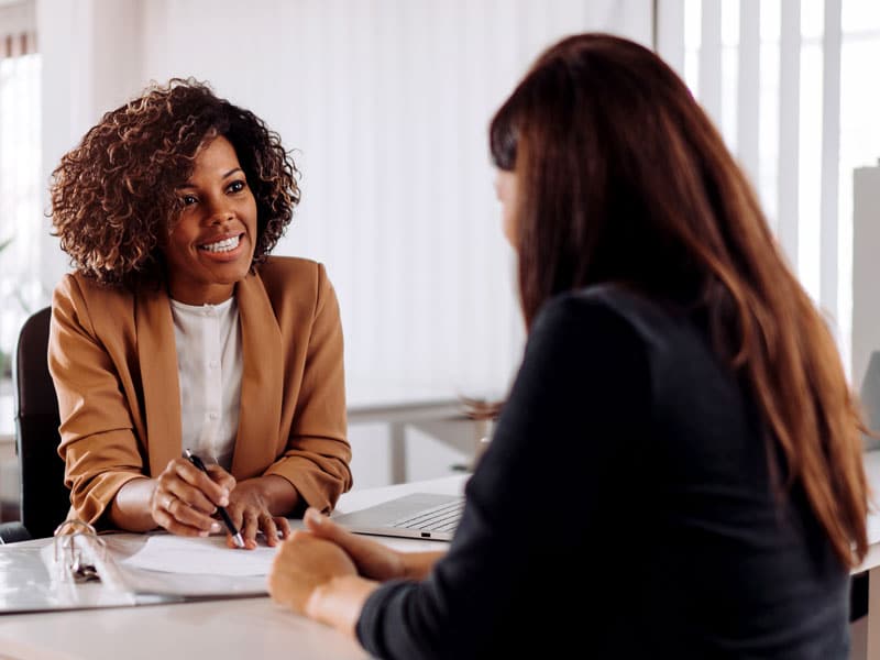 Two women sit across from each other at an office desk; one smiles and writes on papers while the other listens attentively.
